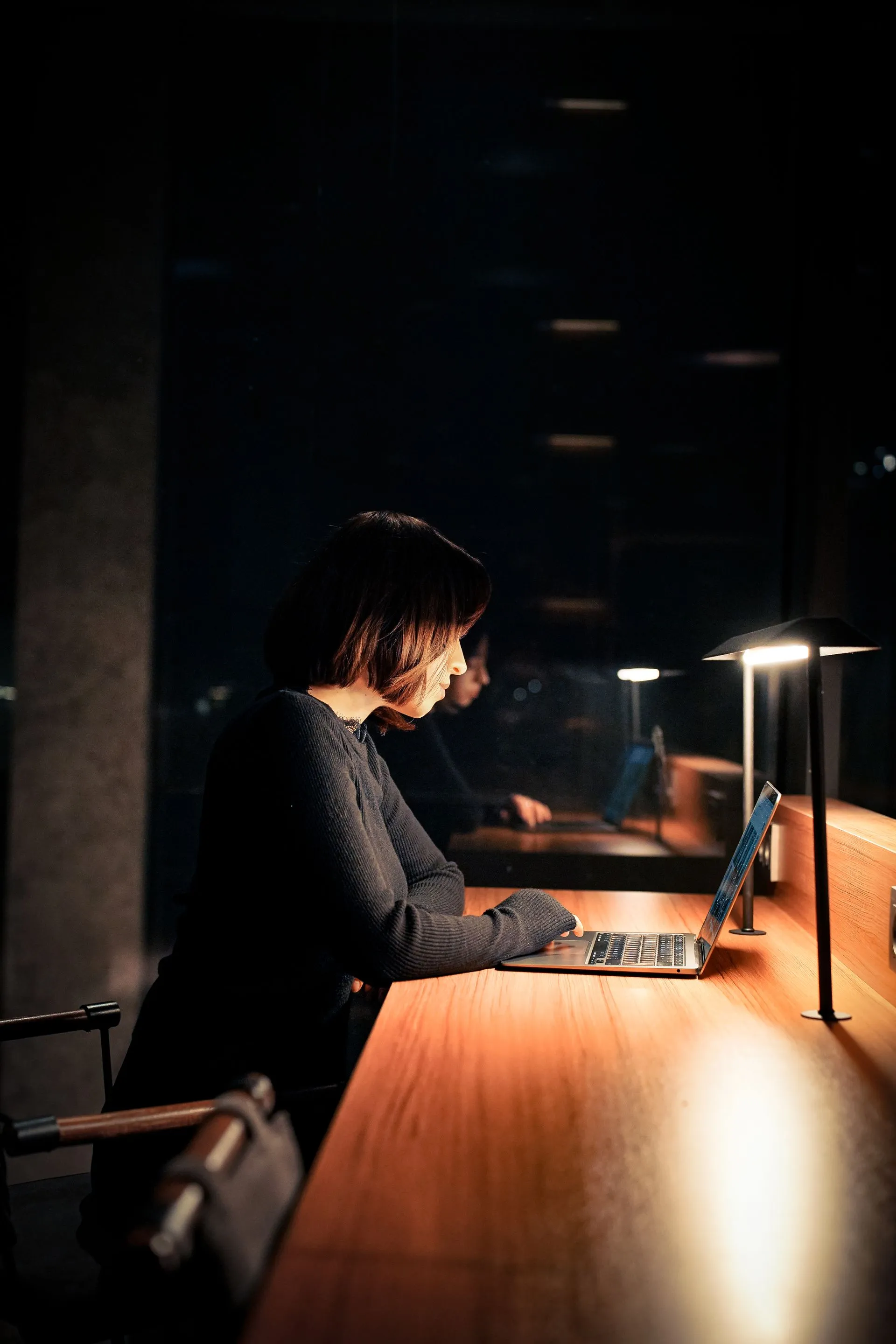 A person working alone at her laptop in a darkened office, lit only by her desk lamp — the work is real, the room is not.