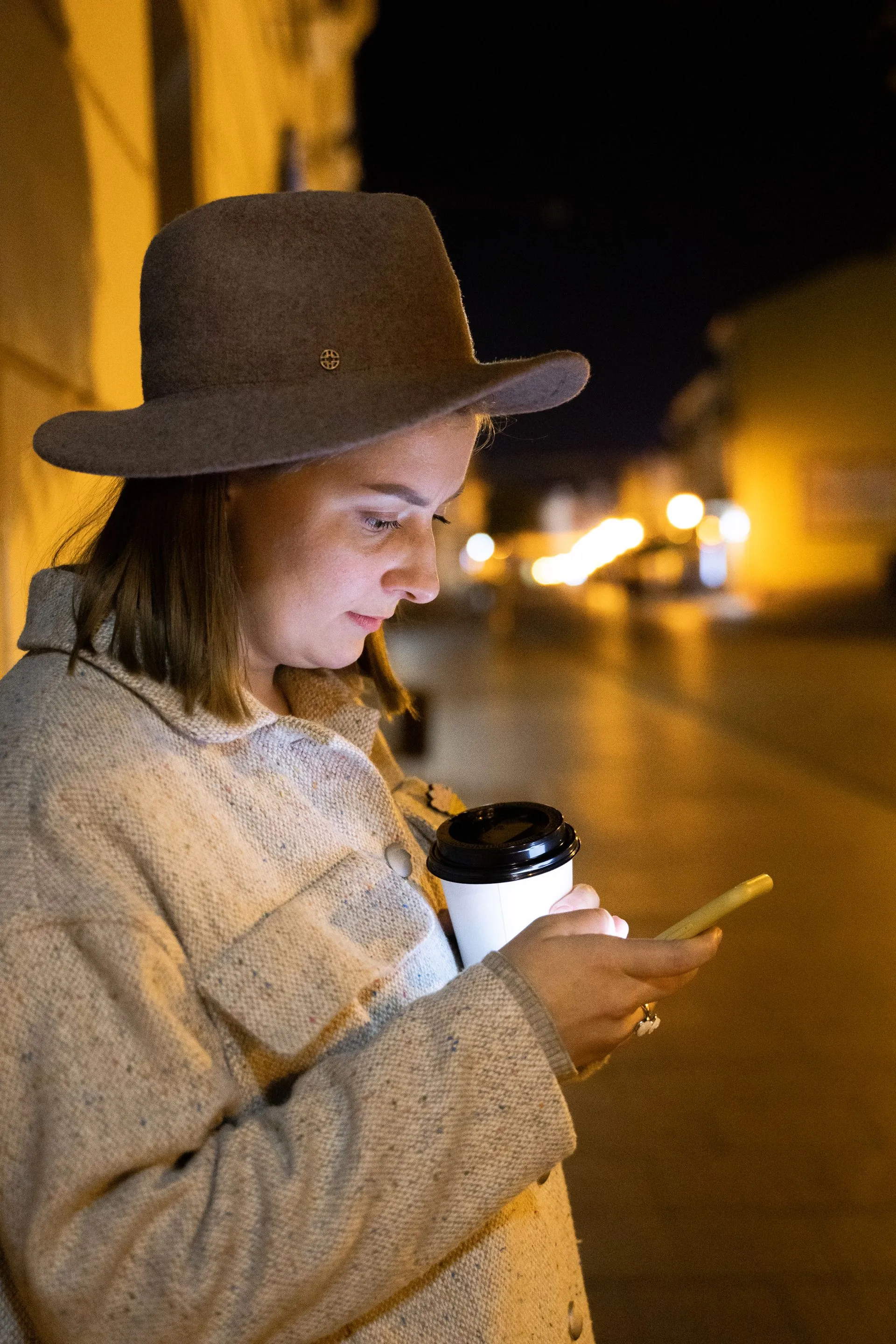 A woman on a night street checking her phone, the screen lighting her hands.